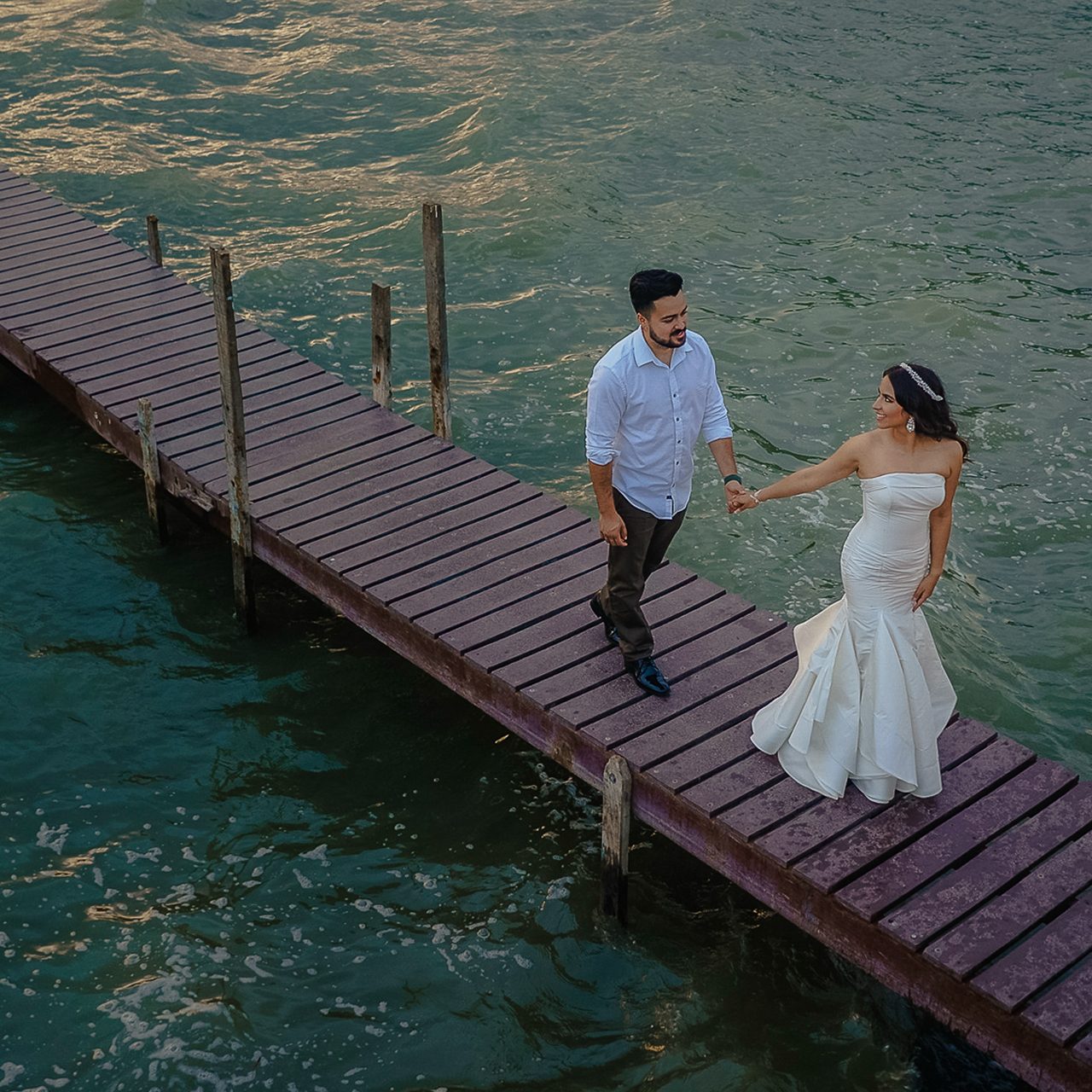 Bride and groom walking hand in hand on a wooden pier by the water, smiling, natural wedding moment